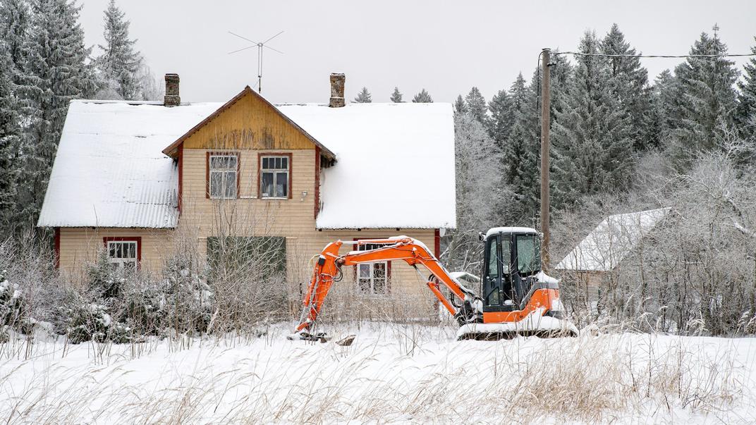 A small orange excavator parked in fresh snow in front of a house.